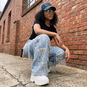 May include: Light blue denim jeans with silver fringe detailing. The jeans have a high waist and a wide leg. The model is wearing a black t-shirt, sunglasses, and a baseball cap. White platform sneakers complete the look.