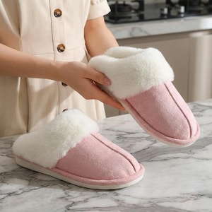 Peut inclure: Une paire de pantoufles roses et blanches en peluche. Les pantoufles ont un ext&eacute;rieur doux, semblable &agrave; du daim, de couleur rose clair, compl&eacute;t&eacute; par une garniture blanche duveteuse autour de l'ouverture. Les semelles sont blanches.
