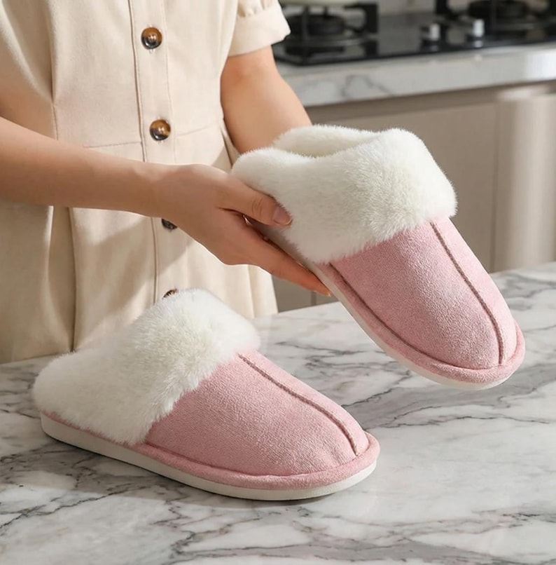 Peut inclure: Une paire de pantoufles roses et blanches en peluche. Les pantoufles ont un ext&eacute;rieur doux, semblable &agrave; du daim, de couleur rose clair, compl&eacute;t&eacute; par une garniture blanche duveteuse autour de l'ouverture. Les semelles sont blanches.