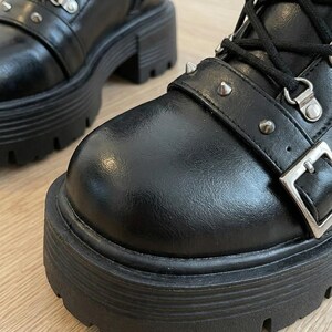 May include: Black leather combat boots with a chunky platform sole. The boots feature silver-tone metal hardware, including eyelets, buckles, and decorative studs. The boots have a lace-up closure and a side zipper.