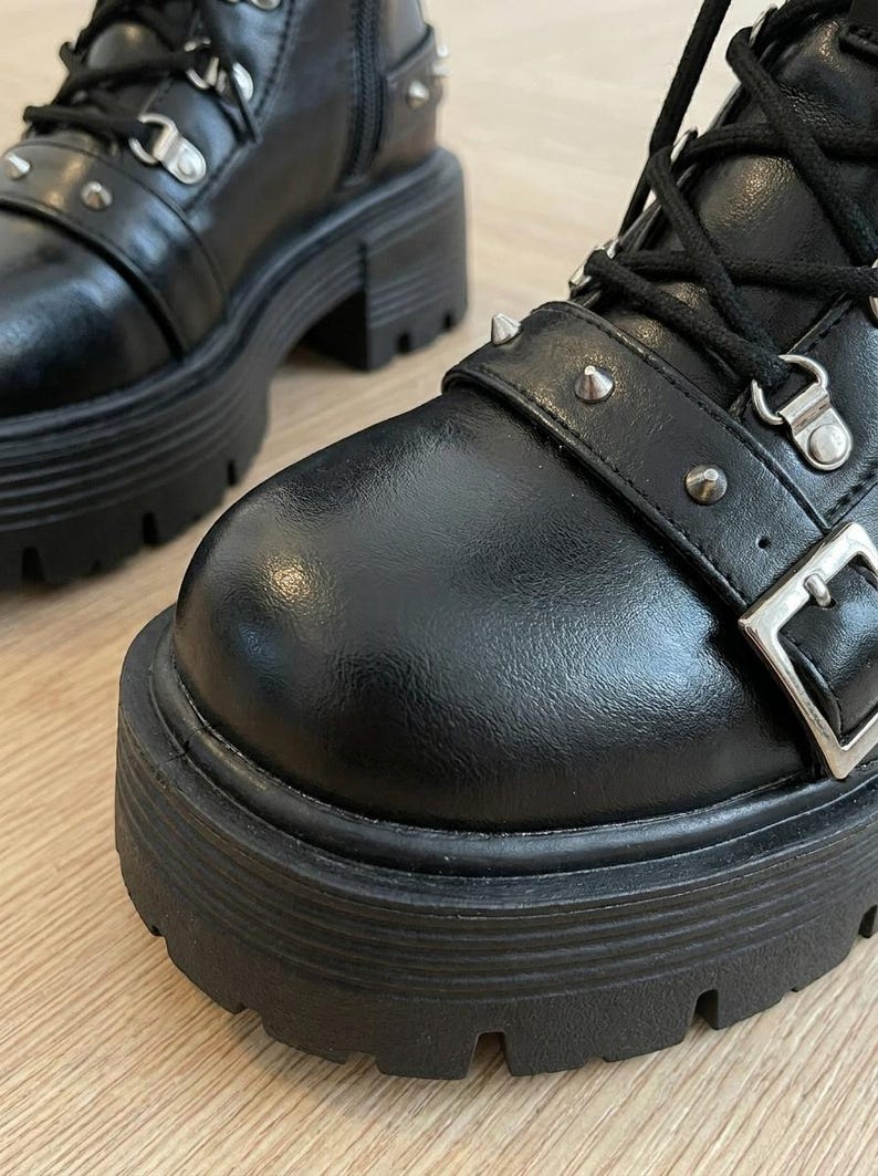 May include: Black leather combat boots with a chunky platform sole. The boots feature silver-tone metal hardware, including eyelets, buckles, and decorative studs. The boots have a lace-up closure and a side zipper.