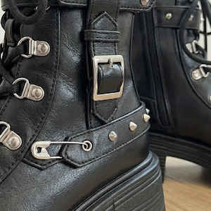 May include: Black leather ankle boots with silver-tone hardware, including buckles, rivets, and safety pins. The boots have a chunky platform sole and lace-up closure, showcasing a punk-inspired design.