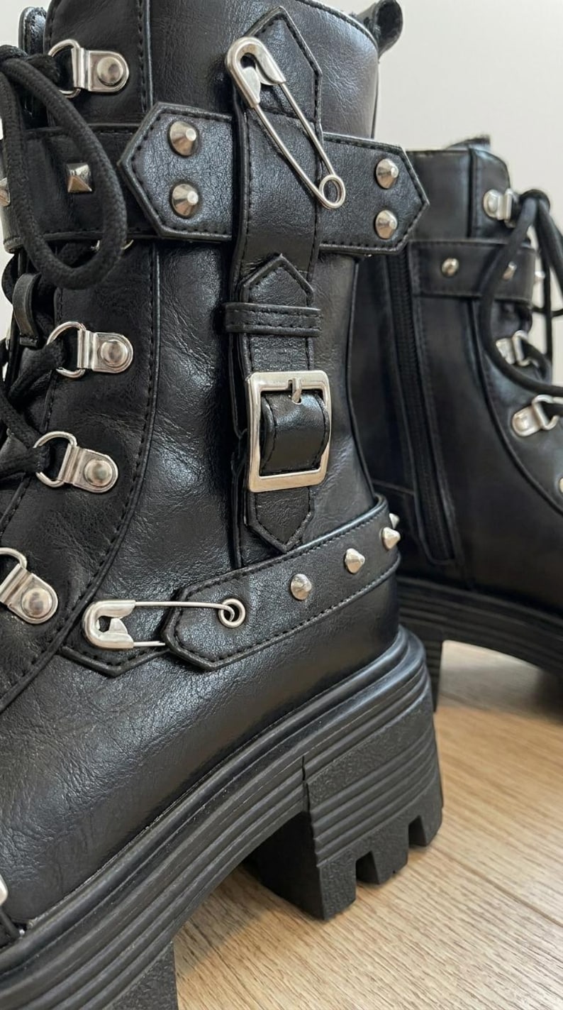 May include: Black leather ankle boots with silver-tone hardware, including buckles, rivets, and safety pins. The boots have a chunky platform sole and lace-up closure, showcasing a punk-inspired design.