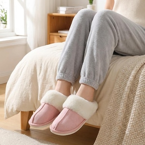 Peut inclure: Une paire de pantoufles roses en daim avec une bordure en fausse fourrure blanche. Les pantoufles sont sur un lit avec une couette beige. La personne porte un pantalon de surv&ecirc;tement gris. Ces chaussons d'int&eacute;rieur offrent confort et chaleur.