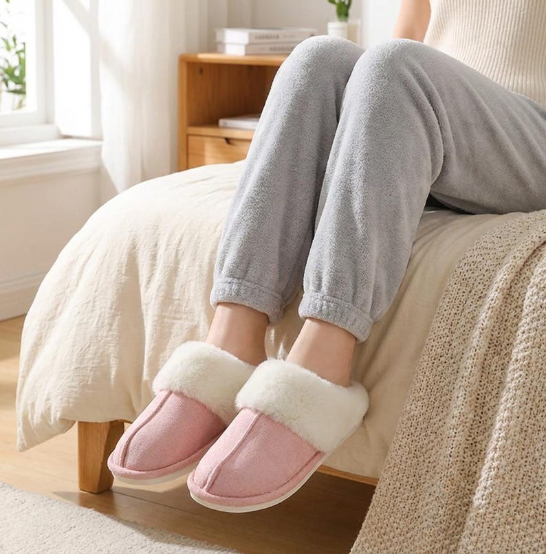 Peut inclure: Une paire de pantoufles roses en daim avec une bordure en fausse fourrure blanche. Les pantoufles sont sur un lit avec une couette beige. La personne porte un pantalon de surv&ecirc;tement gris. Ces chaussons d'int&eacute;rieur offrent confort et chaleur.