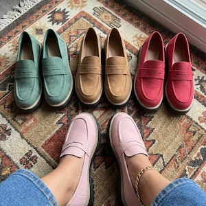May include: A selection of penny loafers in shades of green, tan, red, and pink. The shoes are displayed on a patterned rug, highlighting their classic design and casual style. The pink loafers are being worn.