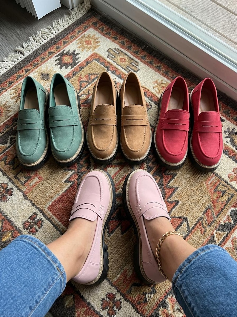 May include: A selection of penny loafers in shades of green, tan, red, and pink. The shoes are displayed on a patterned rug, highlighting their classic design and casual style. The pink loafers are being worn.