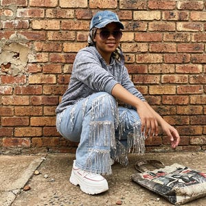 May include: Light blue denim jeans with silver fringe detailing. The jeans are paired with white sneakers with red accents. A gray hooded sweatshirt and a denim baseball cap complete the casual outfit. A printed tote bag rests on the ground.