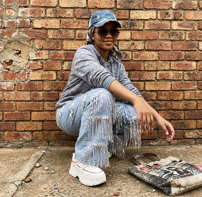 May include: Light blue denim jeans with silver fringe detailing. The jeans are paired with white sneakers with red accents. A gray hooded sweatshirt and a denim baseball cap complete the casual outfit. A printed tote bag rests on the ground.