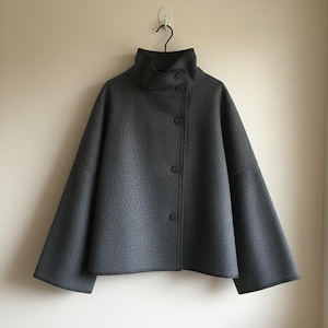 May include: A gray, button-up jacket with a high, rounded collar. The jacket has a textured surface and wide sleeves. The buttons are dark gray, and the jacket is hanging on a black hanger against a neutral wall.