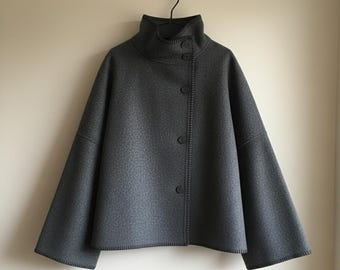 May include: A gray, button-up jacket with a high, rounded collar. The jacket has a textured surface and wide sleeves. The buttons are dark gray, and the jacket is hanging on a black hanger against a neutral wall.