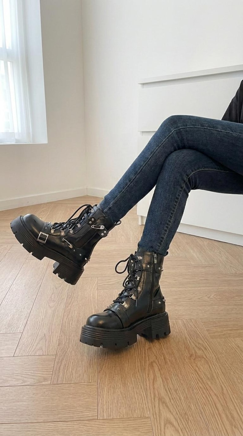 May include: Black leather combat boots with silver-tone hardware, including buckles and studs. The boots have thick, lug soles and lace-up closures. The boots are worn with dark blue denim jeans.