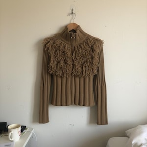 May include: A tan-colored zip-up sweater with a high collar and fringed detailing across the shoulders. The sweater has a ribbed knit design and long sleeves. The zipper has a gold-colored ring pull.