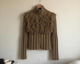 May include: A tan-colored zip-up sweater with a high collar and fringed detailing across the shoulders. The sweater has a ribbed knit design and long sleeves. The zipper has a gold-colored ring pull.