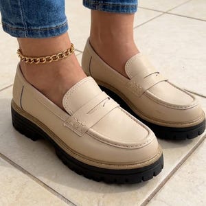 May include: Beige leather loafers with a black sole. The shoes feature a classic penny loafer design with a strap across the vamp. The shoes are worn with blue jeans and a gold chain ankle bracelet.