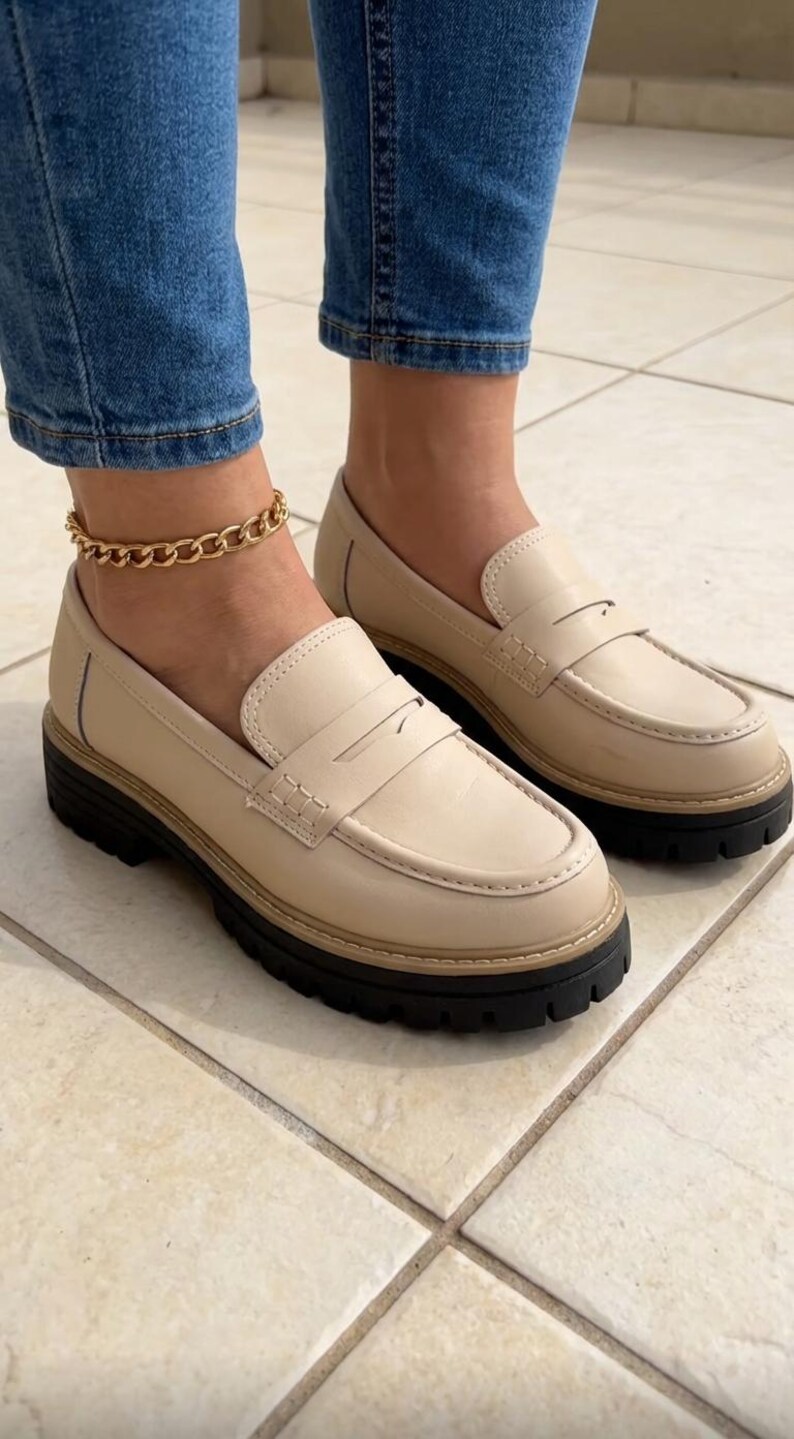 May include: Beige leather loafers with a black sole. The shoes feature a classic penny loafer design with a strap across the vamp. The shoes are worn with blue jeans and a gold chain ankle bracelet.