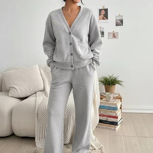 May include: A light grey lounge set comprising a button-down cardigan with pockets and wide-leg sweatpants. The model wears white trainers. The outfit is made of a soft, comfortable material. The set is ideal for casual wear.