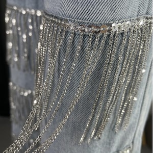 May include: Close-up of light blue denim jeans with silver chain fringe detailing. The fringe is attached to the jeans with a row of silver sequins. The chains hang vertically, creating a shimmering effect.