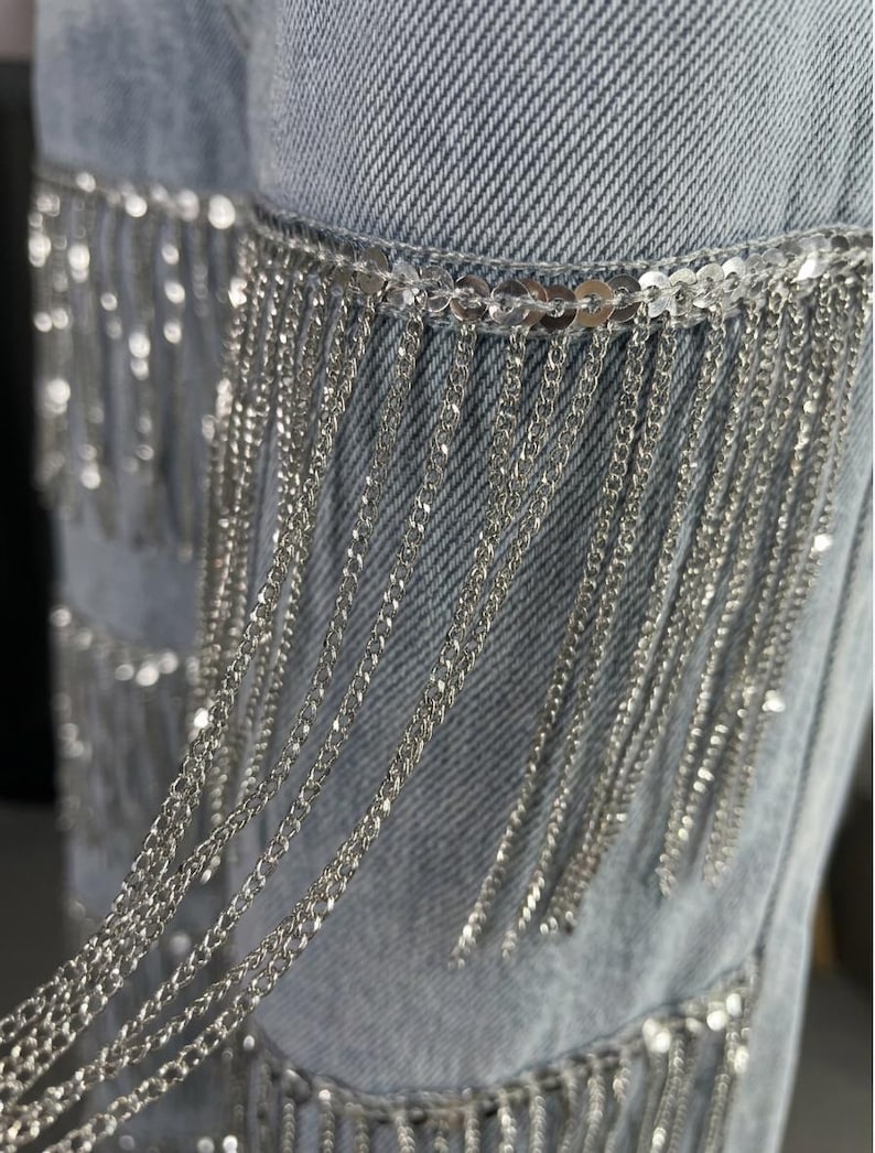 May include: Close-up of light blue denim jeans with silver chain fringe detailing. The fringe is attached to the jeans with a row of silver sequins. The chains hang vertically, creating a shimmering effect.