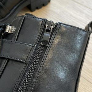 May include: Close-up of a black leather boot with a zipper closure. The boot features a chunky sole, a buckle detail, and a lace-up design. The leather has a smooth texture, and the zipper is metallic.