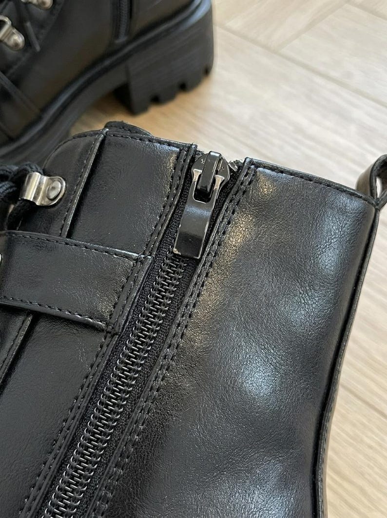 May include: Close-up of a black leather boot with a zipper closure. The boot features a chunky sole, a buckle detail, and a lace-up design. The leather has a smooth texture, and the zipper is metallic.