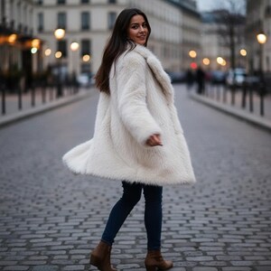 May include: A woman wearing a long, off-white faux fur coat, dark blue jeans, and brown ankle boots. The coat has a wide collar and a flowing silhouette. The background shows a cobblestone street and city buildings.