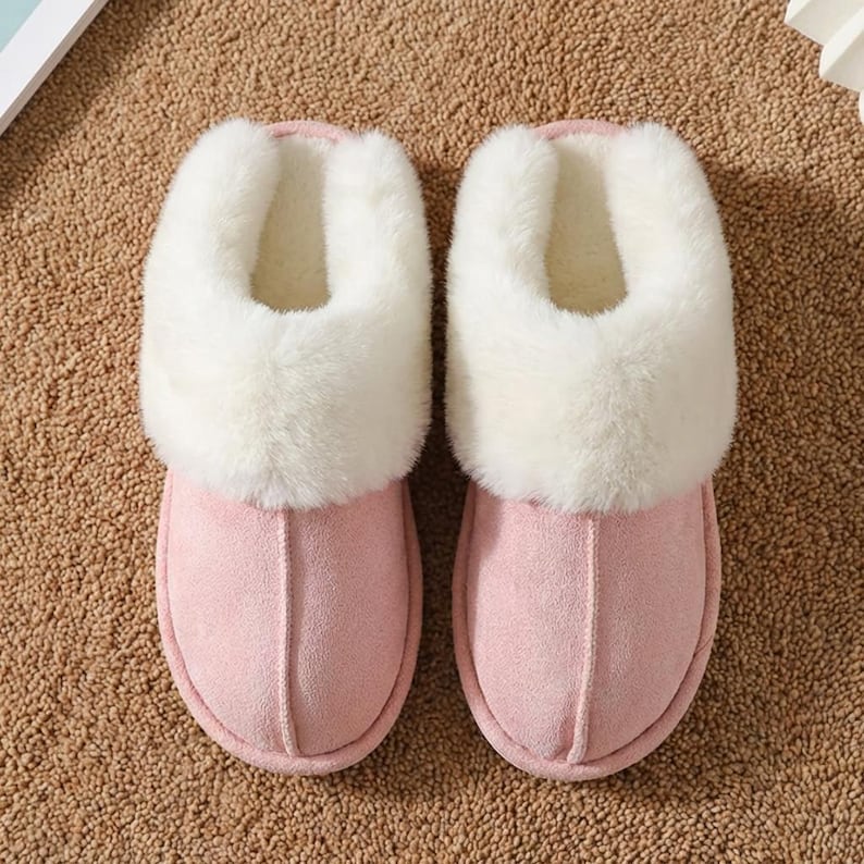 Peut inclure: Une paire de pantoufles roses avec une bordure en fausse fourrure douce. Les pantoufles ont un design ferm&eacute; et sont faites d'un mat&eacute;riau semblable &agrave; du daim. L'int&eacute;rieur est doubl&eacute; d'une mati&egrave;re moelleuse. Pantoufles pour l'int&eacute;rieur.