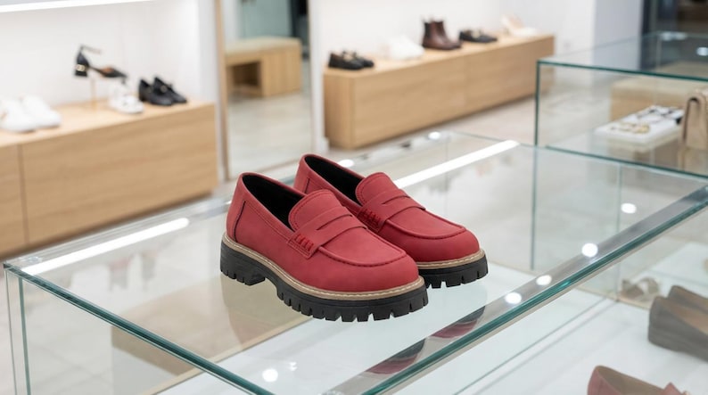 May include: Red leather loafers with a black lug sole, displayed on a glass surface. The shoes have a classic penny loafer design. Other shoes are visible in the background, suggesting a shoe shop setting.