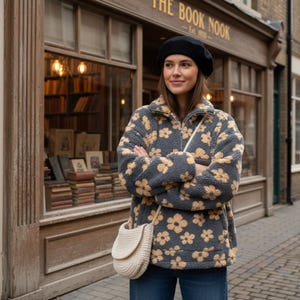Puede incluir: Una mujer con una chaqueta de forro polar gris con estampado floral amarillo, una boina negra y un bolso cruzado color crema. El fondo presenta una librería con el letrero "The Book Nook".