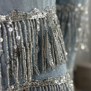 May include: Close-up of light blue denim fabric embellished with silver sequins and fringe. The silver fringe hangs vertically, creating a layered effect. The sequins are sewn along the edges and in rows, adding sparkle.