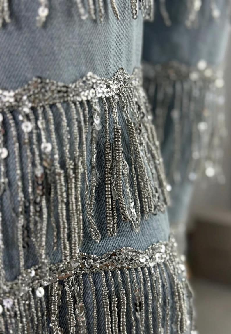 May include: Close-up of light blue denim fabric embellished with silver sequins and fringe. The silver fringe hangs vertically, creating a layered effect. The sequins are sewn along the edges and in rows, adding sparkle.