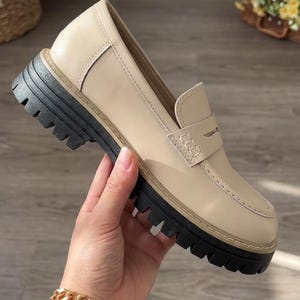 May include: A beige leather loafer with a thick black sole. The shoe has a classic penny loafer design with a stitched vamp and a rounded toe. The shoe is being held by a person.