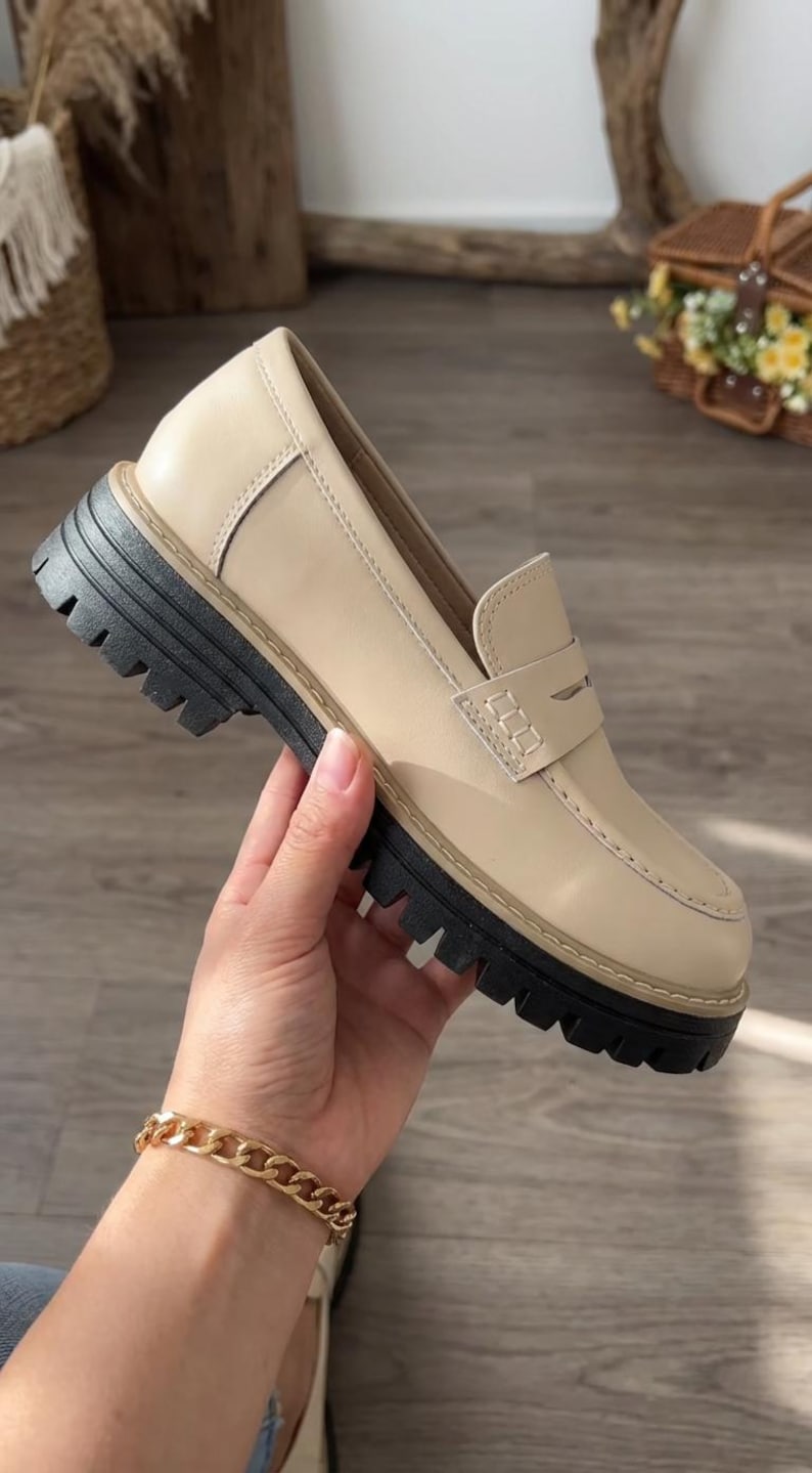 May include: A beige leather loafer with a thick black sole. The shoe has a classic penny loafer design with a stitched vamp and a rounded toe. The shoe is being held by a person.