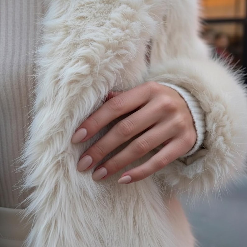 May include: A close-up of a hand with light beige nail polish, resting on a fluffy, off-white faux fur coat. The coat has a soft, textured appearance, and the sleeve of a white sweater is visible.