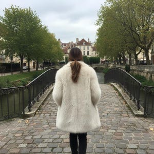 May include: A person wearing a cream-colored faux fur coat, standing on a cobblestone bridge. The coat has a fluffy texture and a mid-length cut. The background features a river, trees, and buildings.