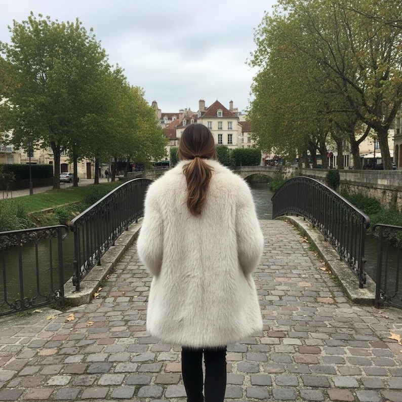 May include: A person wearing a cream-colored faux fur coat, standing on a cobblestone bridge. The coat has a fluffy texture and a mid-length cut. The background features a river, trees, and buildings.