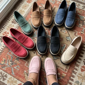 May include: A selection of penny loafers in shades of green, tan, blue, red, black, pink, and cream. The shoes are displayed on a patterned rug, highlighting their diverse colours and designs. The loafers feature a traditional design.