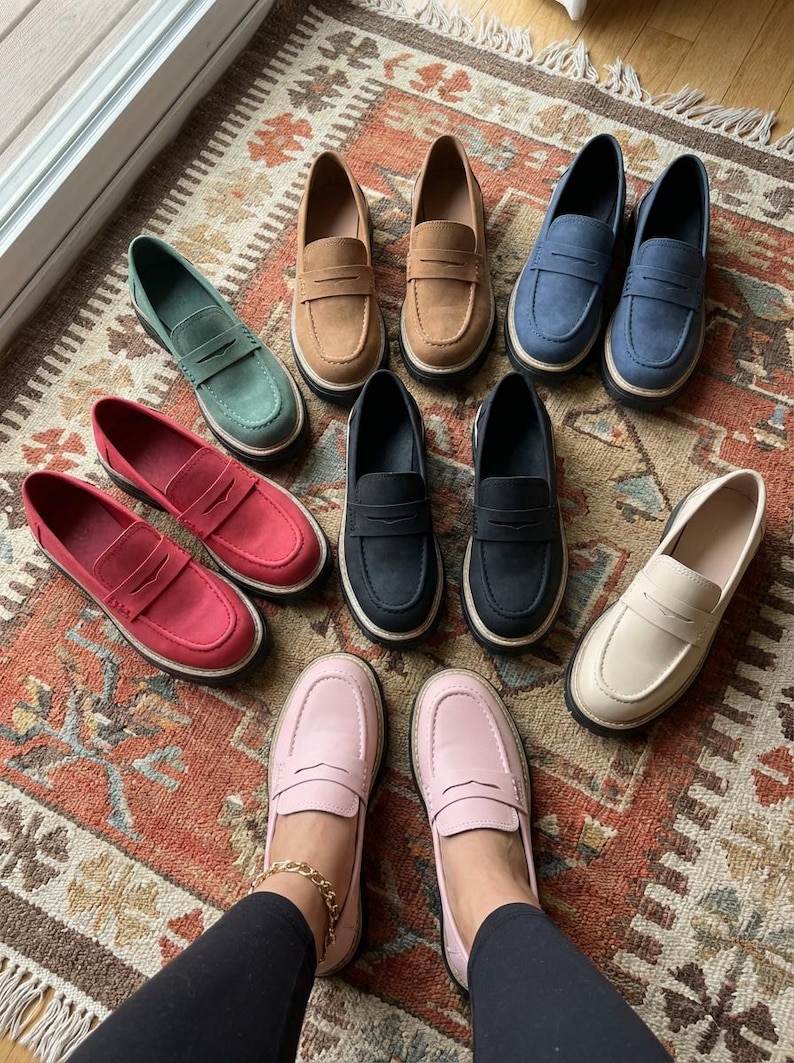 May include: A selection of penny loafers in shades of green, tan, blue, red, black, pink, and cream. The shoes are displayed on a patterned rug, highlighting their diverse colours and designs. The loafers feature a traditional design.