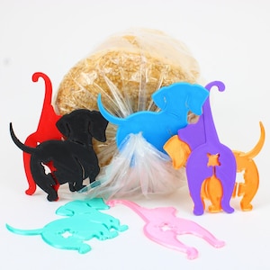 May include: A collection of colorful, dog-shaped bag clips in red, black, blue, purple, orange, pink, and teal. The clips are made of plastic and are designed to seal bags of food. A loaf of bread is visible in the background.