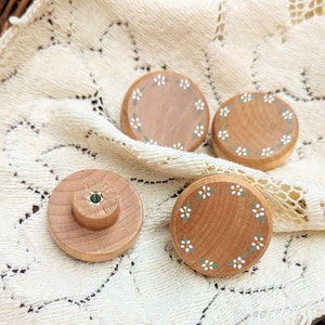 May include: Four wooden cabinet knobs and drawer pulls. Three round knobs feature a painted floral design with white flowers and green leaves. One knob has a small, round handle with a green center. The knobs are made of light-colored wood.