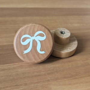May include: Two wooden cabinet knobs. One knob features a light blue bow design on a round, light brown surface. The second knob is light brown with a cylindrical base and a round top. The knobs are made of wood.