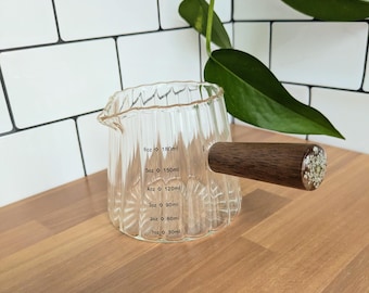 May include: A clear, ribbed glass measuring pitcher with a spout and a dark brown wooden handle. The handle features a circular end cap containing embedded white dried flowers. Measurement markings range from 1oz to 6oz and 30ml to 180ml.