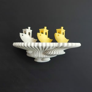 May include: Three miniature boat sculptures in white and yellow rest on a decorative shelf. The shelf has a unique, wavy design with white and gray stripes. The background is a solid black, creating a contrast that highlights the objects.