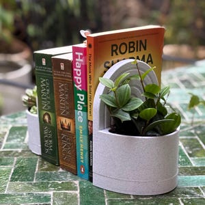Peut inclure: Un pot décoratif blanc incurvé contenant une petite plante et plusieurs livres, dont "Happy Place" et "The Monk Who Sold His Ferrari". Le pot est posé sur une surface carrelée verte. Les livres sont alignés.