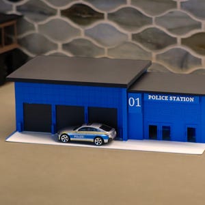 Könnte beinhalten: Ein blaues Modell einer Polizeistation mit schwarzem Flachdach, drei Garagentoren und dem Aufdruck "01" sowie "POLICE STATION" an der Vorderseite. Ein kleines silbernes Spielzeugauto mit der Aufschrift "POLIZEI" steht vor einem der Garagentore.