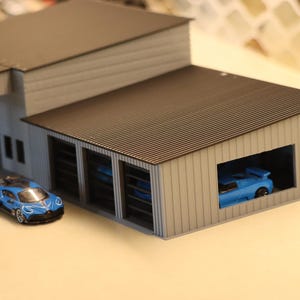 May include: A miniature model of a car garage with a blue sports car parked inside and another one parked outside. The garage has a brown corrugated roof and white siding. The car outside is blue with black accents.