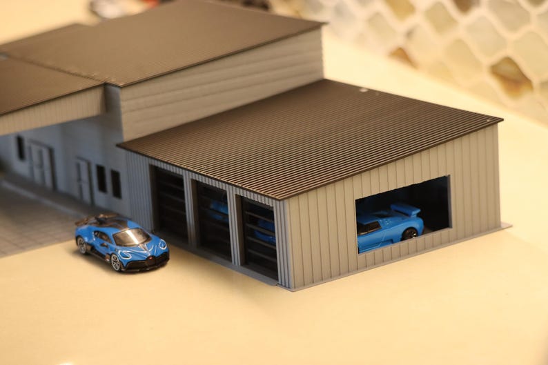 May include: A miniature model of a car garage with a blue sports car parked inside and another one parked outside. The garage has a brown corrugated roof and white siding. The car outside is blue with black accents.