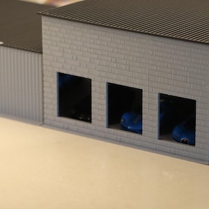 May include: A miniature model of a garage or industrial building. The structure has a gray brick facade with three open bays, each housing a small blue car. The roof is dark gray, and the side walls are corrugated.