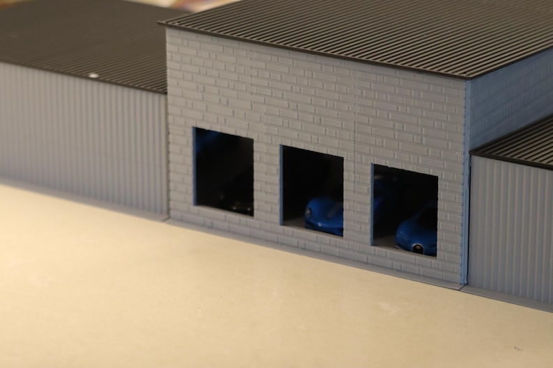 May include: A miniature model of a garage or industrial building. The structure has a gray brick facade with three open bays, each housing a small blue car. The roof is dark gray, and the side walls are corrugated.