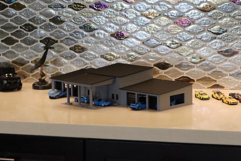 May include: A miniature model of a modern garage with a flat roof and multiple bays, housing small blue sports cars. Several yellow and black toy cars are parked nearby. A black Jeep and a decorative sculpture are also present.